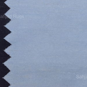 May include: A close-up of a blue rug with a black zigzag border. The rug has a textured appearance and the words "Sahjad Rugs" are printed on the rug.