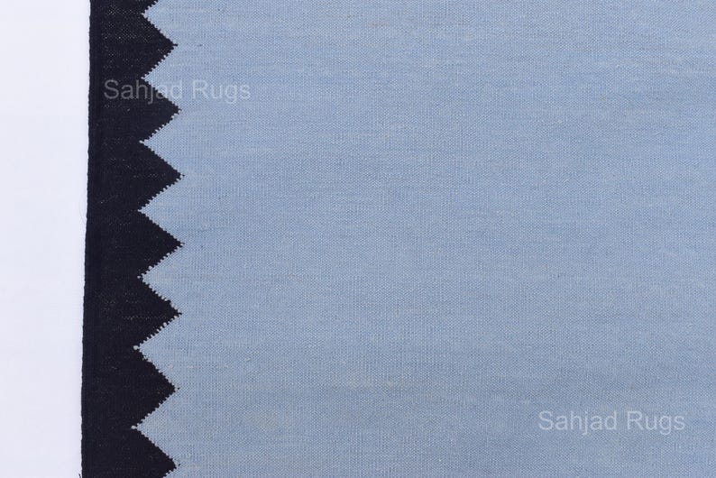 May include: A close-up of a blue rug with a black zigzag border. The rug has a textured appearance and the words "Sahjad Rugs" are printed on the rug.