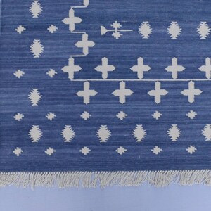 Puede incluir: Una alfombra azul y blanca con un borde de flecos. La alfombra tiene un dise&ntilde;o geom&eacute;trico con cruces y diamantes blancos sobre un fondo azul.