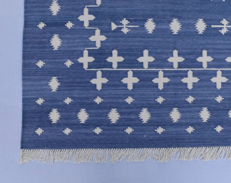 Puede incluir: Una alfombra azul y blanca con un borde de flecos. La alfombra tiene un dise&ntilde;o geom&eacute;trico con cruces y diamantes blancos sobre un fondo azul.
