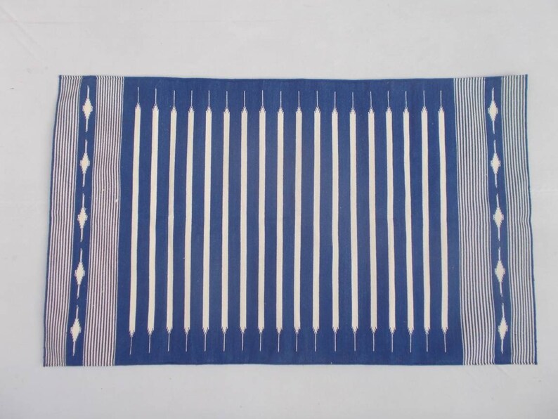 Peut inclure: Un tapis ray&eacute; bleu et blanc avec un motif g&eacute;om&eacute;trique aux extr&eacute;mit&eacute;s. Le tapis est fait d'un mat&eacute;riau tiss&eacute; et a une bordure frang&eacute;e.