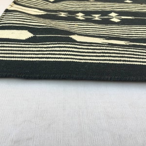 K&ouml;nnte beinhalten: Ein schwarz-cremefarbener gewebter Textil mit einem geometrischen Muster. Das Design zeigt horizontale Streifen und Rauten. Die Kanten des Textils sind sichtbar und zeigen die gewebte Textur.