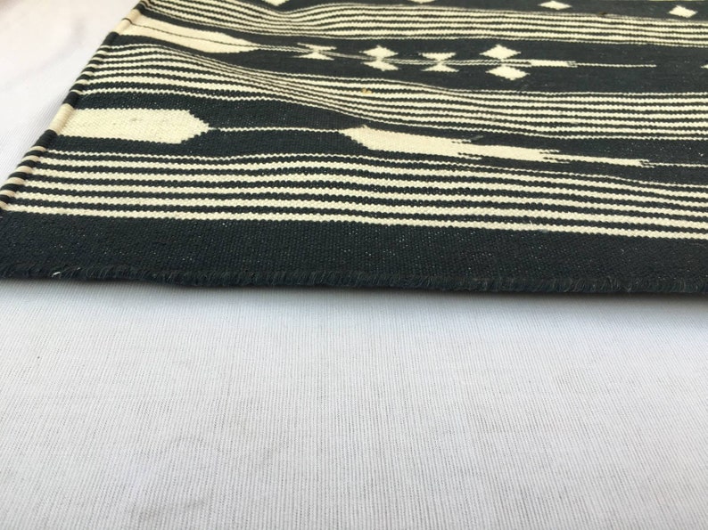 K&ouml;nnte beinhalten: Ein schwarz-cremefarbener gewebter Textil mit einem geometrischen Muster. Das Design zeigt horizontale Streifen und Rauten. Die Kanten des Textils sind sichtbar und zeigen die gewebte Textur.