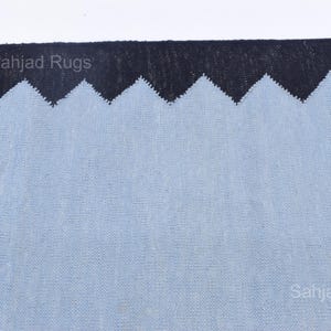 May include: A close-up of a rug with a light blue field and a black zigzag border. The rug has a textured appearance. The text "Sahjad Rugs" is visible in the image.