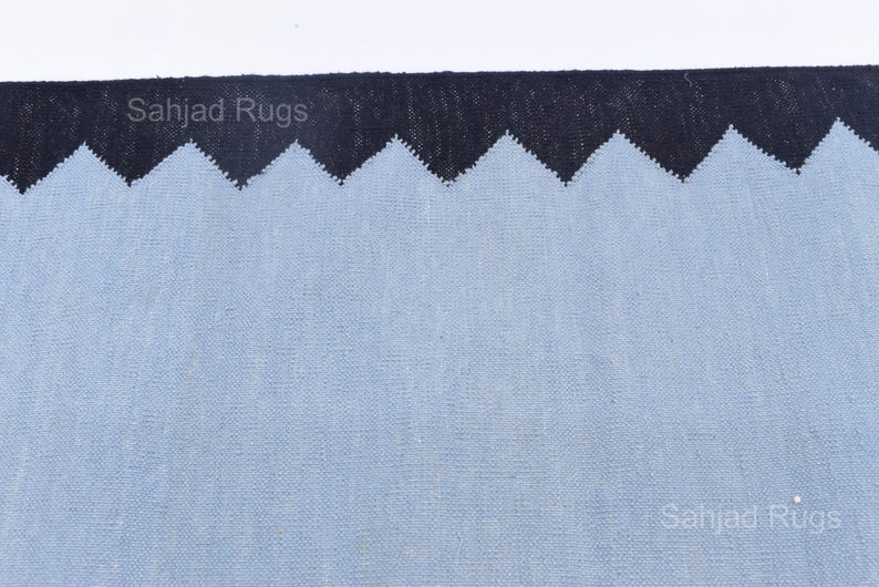 May include: A close-up of a rug with a light blue field and a black zigzag border. The rug has a textured appearance. The text "Sahjad Rugs" is visible in the image.