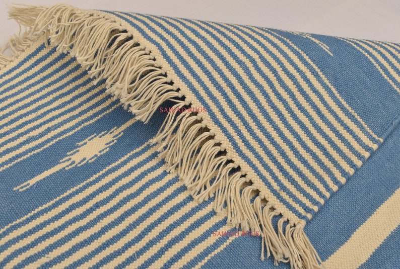 May include: Blue and cream striped rug with fringed edges. The rug features a repeating pattern of diagonal stripes in light blue and cream. The fringed edge adds a decorative element to the rug.