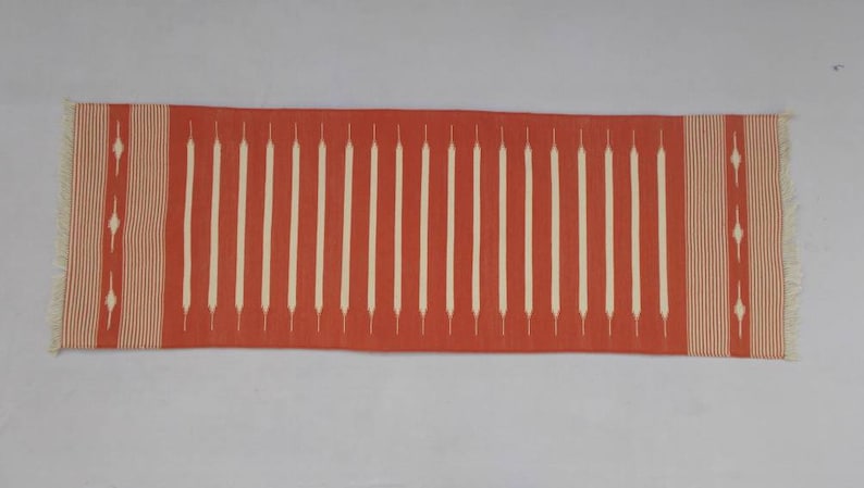 May include: A red and white runner rug with a geometric pattern. The rug has a fringe on both ends.