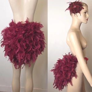 May include: A burgundy feather skirt and matching headpiece. The skirt is made of dense, fluffy feathers. The headpiece features a flower design with a central jewel. The outfit is displayed on a mannequin.