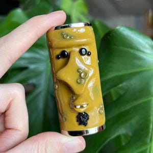 May include: A yellow, cylindrical art piece with a face design. The face has black eyes, a star-shaped nose, and a small black beard. The piece has a silver base and top. The art piece is held by a hand.
