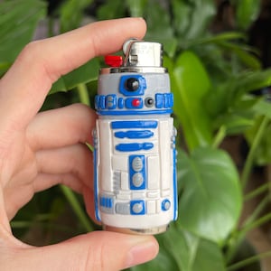 Pode incluir: Um isqueiro R2-D2 branco e azul com uma parte superior prateada. O isqueiro está sendo segurado em uma mão.