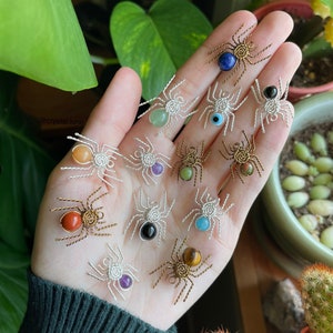 以下が含まれることがあります： 手の中に、中心に異なる色の宝石が埋め込まれた、ワイヤーで巻かれた小さなクモが13匹あります。宝石には、ラピスラズリ、グリーンアベンチュリン、ターコイズ、タイガーアイ、レッドジャスパー、アメジスト、ブラックオニキスが含まれています。