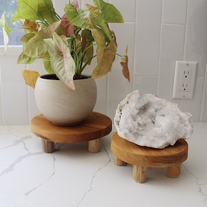 May include: Two wooden plant stands with three legs each. The stand on the left has a white ceramic pot with a leafy green plant. The stand on the right has a large white crystal on top.