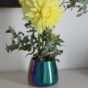 May include: A vibrant floral arrangement featuring large yellow dahlias and eucalyptus leaves in a metallic iridescent vase. The vase has a smooth, reflective surface with a blend of blue, green, and purple hues. The flowers are the focal point.