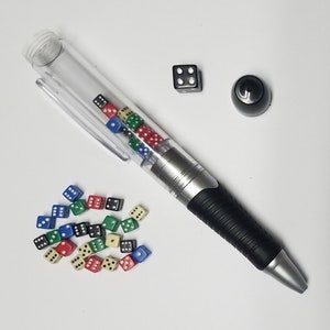 Op de afbeelding: Een transparante pen met een zwarte grip en een zilveren cilinder gevuld met kleurrijke dobbelstenen. De pen ligt op een wit oppervlak met een zwarte dobbelsteen en een zwarte dop.
