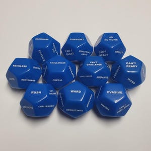May include: A collection of ten blue polyhedral dice with white text. Each die face displays a different action or command, such as "Support," "Rush," and "Evasive." The dice are arranged on a white surface.