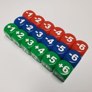 Pode incluir: Um conjunto de dados coloridos, cada um exibindo um número com um sinal de mais ou menos. Os dados são dispostos em fileiras, com dados verdes, azuis e vermelhos. Os números variam de -1 a -6 e +1 a +6. Estes dados são provavelmente usados para jogos de tabuleiro ou fins educacionais.