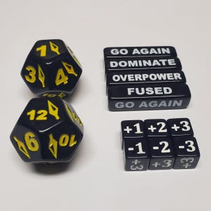 May include: A selection of black gaming dice and tokens. Two large, twelve-sided dice with yellow numerals are shown with black tokens bearing white text, including "GO AGAIN" and "FUSED". Small black dice with white numbers and symbols are also present.