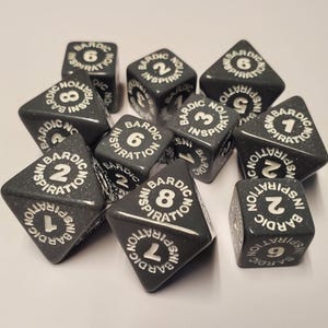 May include: A collection of dark gray, speckled dice with white lettering. Each die features a number from 1 to 8 and the words "BARDIC INSPIRATION" around the edges. The dice are designed for tabletop role-playing games.