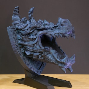 May include: A 3D printed dragon head sculpture with a textured, dark blue finish. The dragon's mouth is open, revealing sharp teeth. The sculpture is mounted on a black stand.