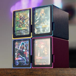 May include: Four card storage boxes in yellow, blue, black, and magenta, each holding a trading card. The cards feature fantasy art and text. The boxes are stacked on top of each other.