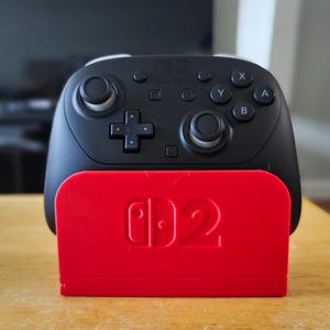 May include: A black Nintendo Switch Pro Controller with a red charging stand. The controller has a D-pad, analog sticks, and action buttons. The red stand features the Nintendo Switch logo.