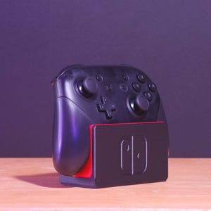 Peut inclure: Une manette Nintendo Switch Pro noire avec un socle de charge rouge et noir. La manette a des boutons étiquetés A, B, X, Y et un pavé directionnel.