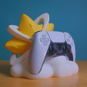 May include: A white and black video game controller is resting on a white cloud-shaped stand with a yellow star on top. The stand is designed to hold the controller upright.