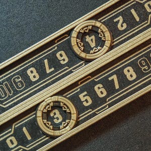 Puede incluir: Primer plano de dos diales num&eacute;ricos negros y dorados con los n&uacute;meros 3 y 4 visibles. Los diales est&aacute;n colocados sobre una superficie negra con n&uacute;meros y l&iacute;neas doradas. El dise&ntilde;o tiene una est&eacute;tica futurista e industrial.