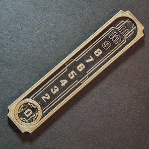 Puede incluir: Un objeto rectangular dorado con un centro negro, con los n&uacute;meros del 2 al 10. El objeto tiene un borde decorativo y un dise&ntilde;o circular en un extremo, con el n&uacute;mero 0. El art&iacute;culo parece ser una pieza decorativa.