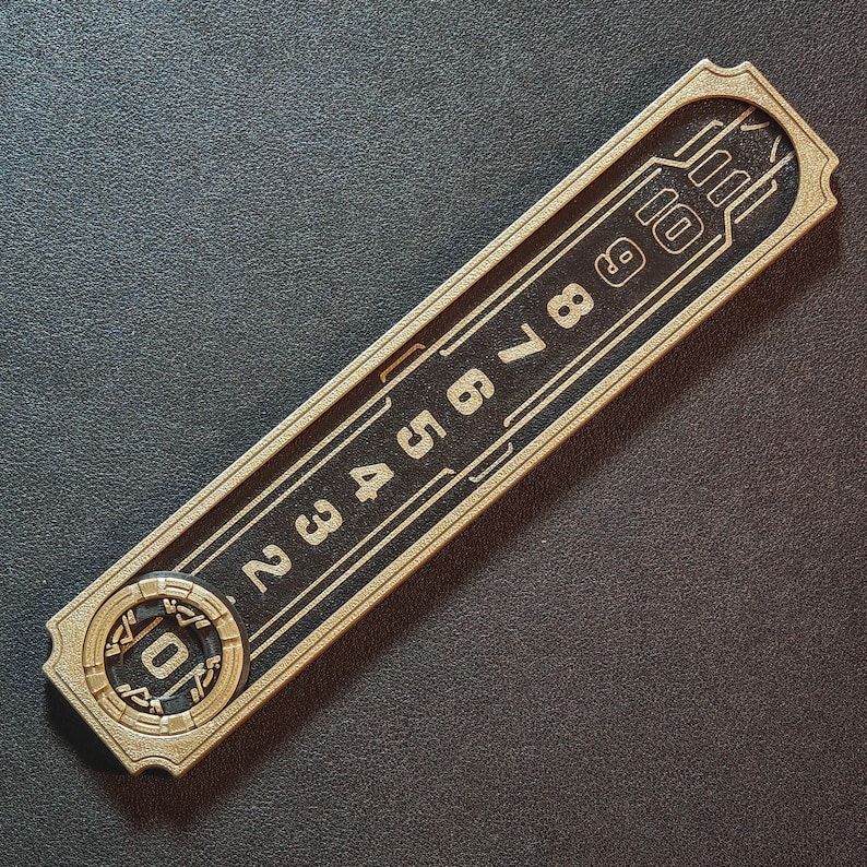Puede incluir: Un objeto rectangular dorado con un centro negro, con los n&uacute;meros del 2 al 10. El objeto tiene un borde decorativo y un dise&ntilde;o circular en un extremo, con el n&uacute;mero 0. El art&iacute;culo parece ser una pieza decorativa.