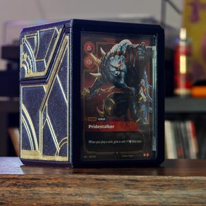 Può includere: Una custodia espositiva rettangolare per carte collezionabili blu scuro e oro con un motivo geometrico ispirato all'Art Déco sul lato. Il pannello frontale trasparente mostra una carta Pridestalker con testo rosso e nero e un'illustrazione.