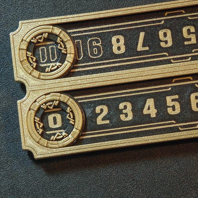 Puede incluir: Dos placas rectangulares negras y doradas con n&uacute;meros dorados en relieve y detalles circulares dorados. La placa superior muestra los n&uacute;meros 567890, y la placa inferior muestra 234560. Los detalles circulares tienen los n&uacute;meros 00 y 0.