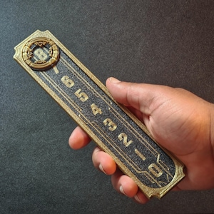 Puede incluir: Panel de ascensor rectangular de estilo Art Déco, con borde dorado y centro negro texturizado. El panel muestra los números del 0 al 8 en dorado, con un diseño circular ornamentado en la parte superior. El panel se sostiene en una mano.