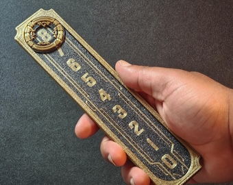 Puede incluir: Panel de ascensor rectangular de estilo Art Déco, con borde dorado y centro negro texturizado. El panel muestra los números del 0 al 8 en dorado, con un diseño circular ornamentado en la parte superior. El panel se sostiene en una mano.