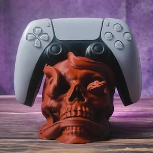 Puede incluir: Un soporte para mando de videojuegos con forma de calavera roja con un mando blanco y negro encima. La calavera tiene detalles intrincados y una superficie texturizada, con una serpiente alrededor. El fondo es morado borroso.