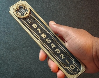 Puede incluir: Un objeto rectangular dorado con detalles negros y números grabados del 0 al 9. El objeto se sostiene en una mano y tiene un diseño circular en la parte superior. Los números están en una línea vertical.