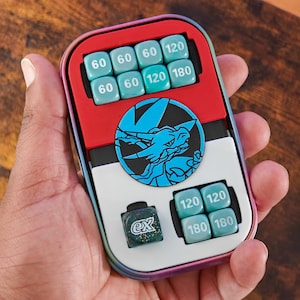 May include: A multi-colored game accessory with a red top, white center, and black and blue accents. It features several teal dice with numbers like 60, 120, and 180. A circular emblem with a blue dragon is visible.