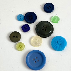 May include: A collection of various colorful buttons in shades of blue, green, and olive. The buttons are round, square, and oval, with two or four holes. Some buttons have a glossy finish, while others are matte.