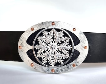 May include: Silver-toned belt buckle with a snowflake design. The oval buckle has a textured silver frame with copper-colored rivets. The snowflake is centered on a black background, attached to a black leather belt.