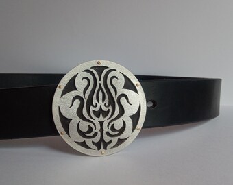 May include: A silver belt buckle with a black floral design. The buckle is round and has a textured surface. The belt is black leather.