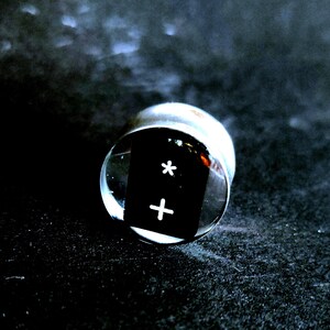 May include: A black and white dice with a plus sign and an asterisk on the faces. The dice is encased in a clear glass sphere.