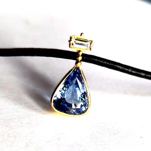 Könnte beinhalten: Goldener Anhänger mit einem hellblauen, birnenförmigen Edelstein. Der Anhänger hängt an einem schwarzen Band. Ein kleinerer, rechteckiger Edelstein ist darüber gesetzt.