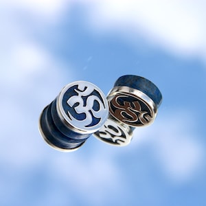 May include: Two silver and blue ear gauges with an Om symbol design. The gauges are made of metal and lapis lazuli.