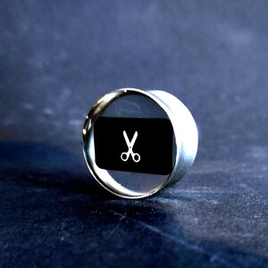 May include: A silver ring with a black background and a white pair of scissors in the center.