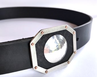 May include: A silver belt buckle with a hammered silver circle in the center, surrounded by a black background and copper accents.