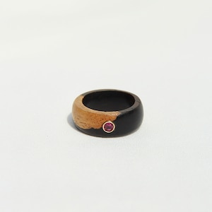 Può includere: Un anello in legno con finitura nera e marrone e una piccola pietra preziosa rosa incastonata al centro.
