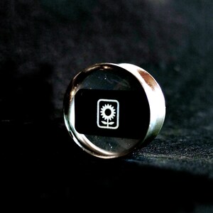 May include: A silver-colored ring with a clear glass top featuring a black background and a white stylized flower design.