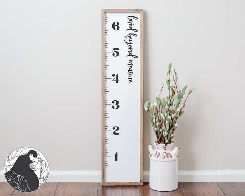 May include: A white wooden growth chart with black numbers 1 through 6 and the text "loved beyond measure" in black cursive. The chart is leaning against a white wall and a white vase with green stems and white flowers is in front of it.