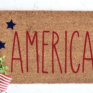 May include: A rectangular doormat with the word "AMERICA" in red, stylized letters. Three dark blue stars are placed around the word. An American flag and greenery are in the lower left corner.