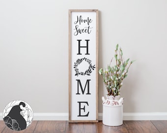 May include: A wooden sign with the words "Home Sweet Home" in black lettering. The letters are large and stylized. The sign is hanging on a white wall with a vase of white flowers in front of it.